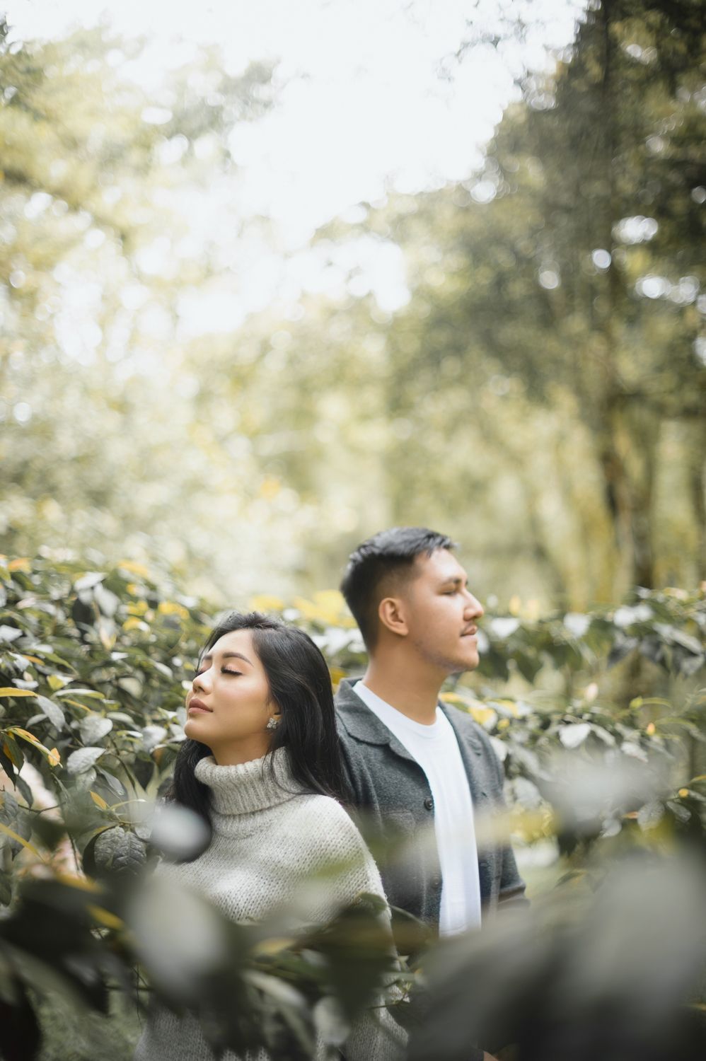 Couple standing among lush green bushes in a sunlit forest, woman in beige turtleneck and man in gray jacket.