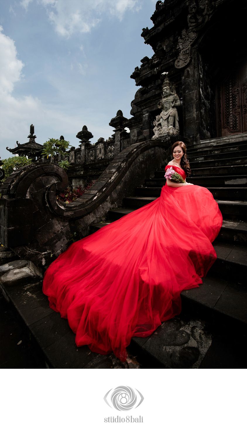 Woman in flowing red gown poses dramatically on ancient Balinese temple steps, dress billowing in the wind.