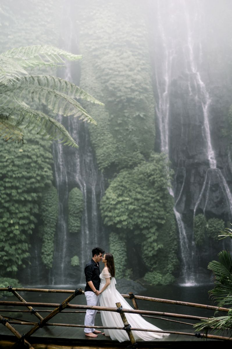 Couple sharing a romantic kiss on a bamboo bridge in front of a lush green waterfall in Bali.