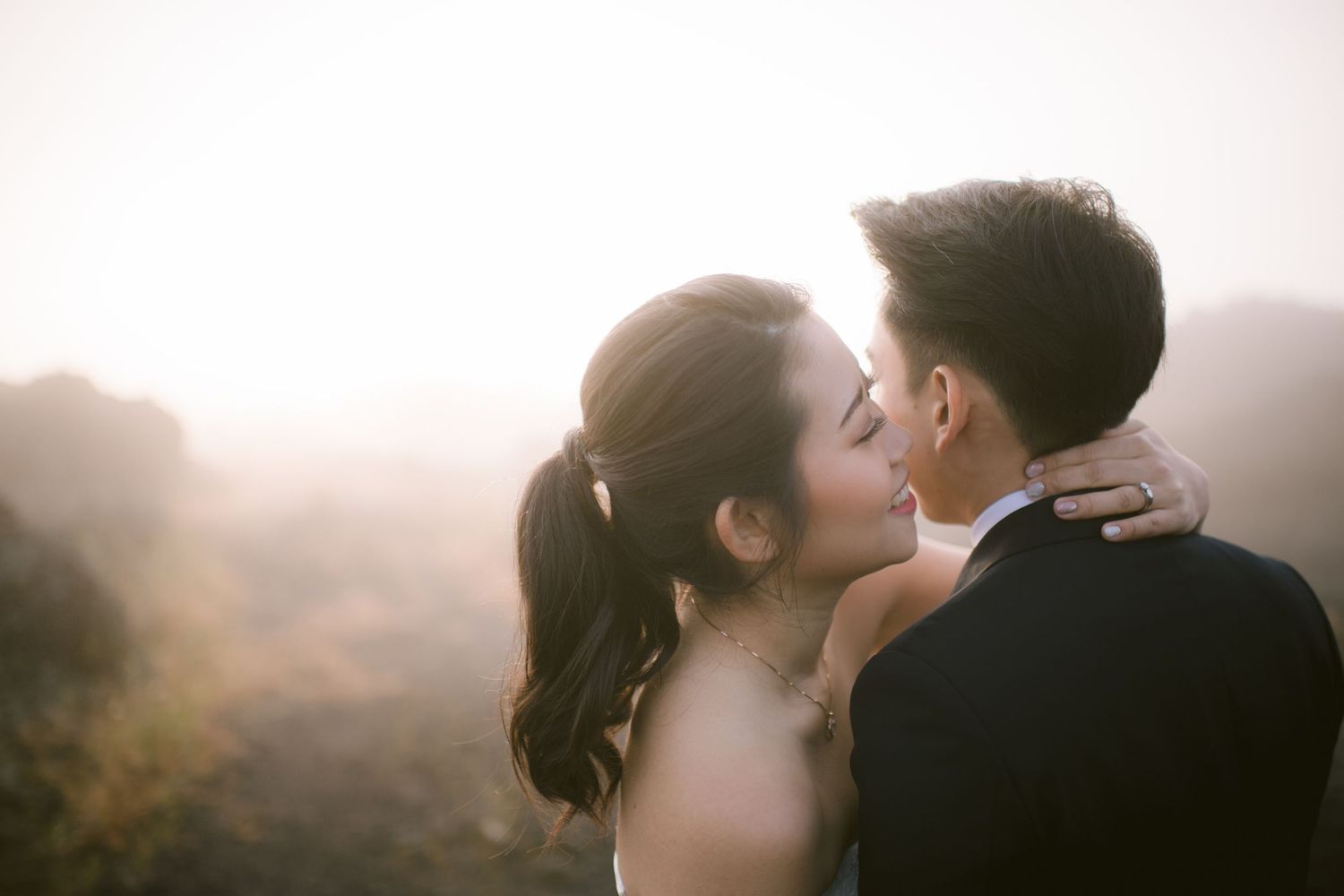 Couple sharing a romantic kiss at golden hour with misty hills in the background at their outdoor wedding.