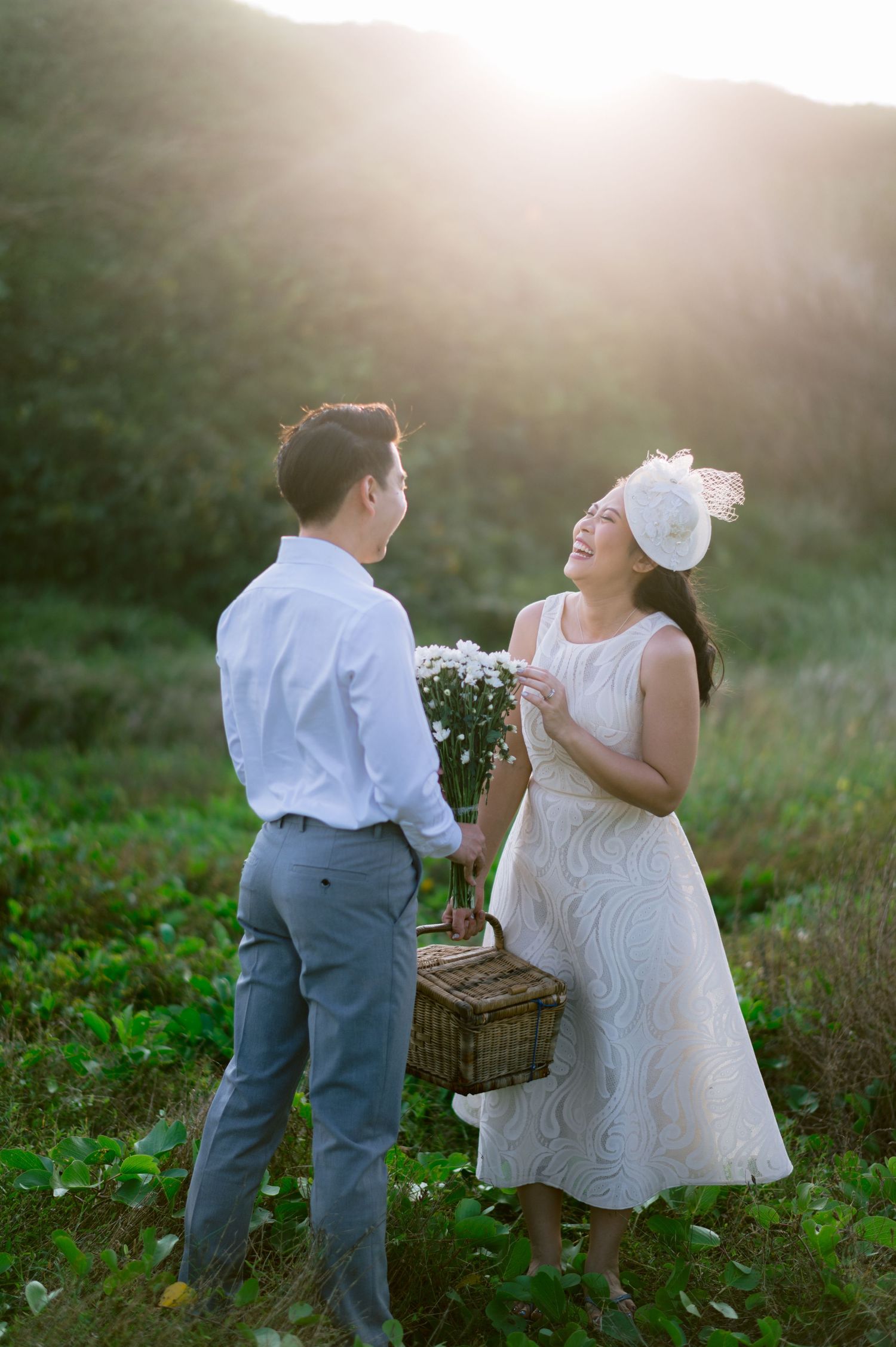 Couple in white attire laughing in a sunlit green field, woman holding flowers and a picnic basket.