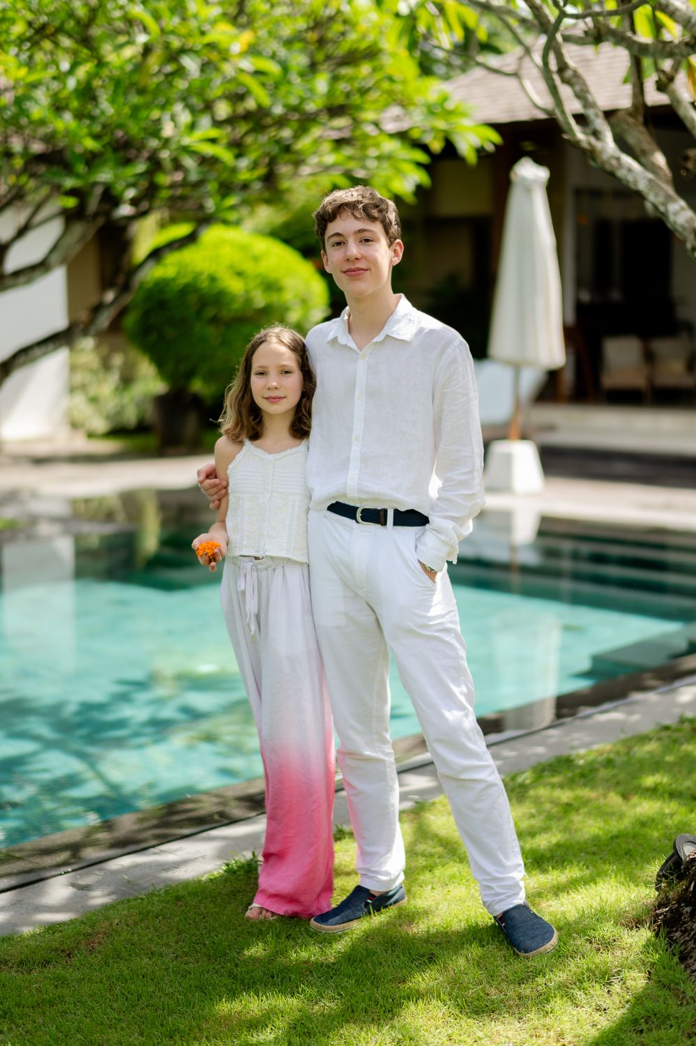 Two children posing together by a pool in a lush garden, dressed in white and pink gradient pants.