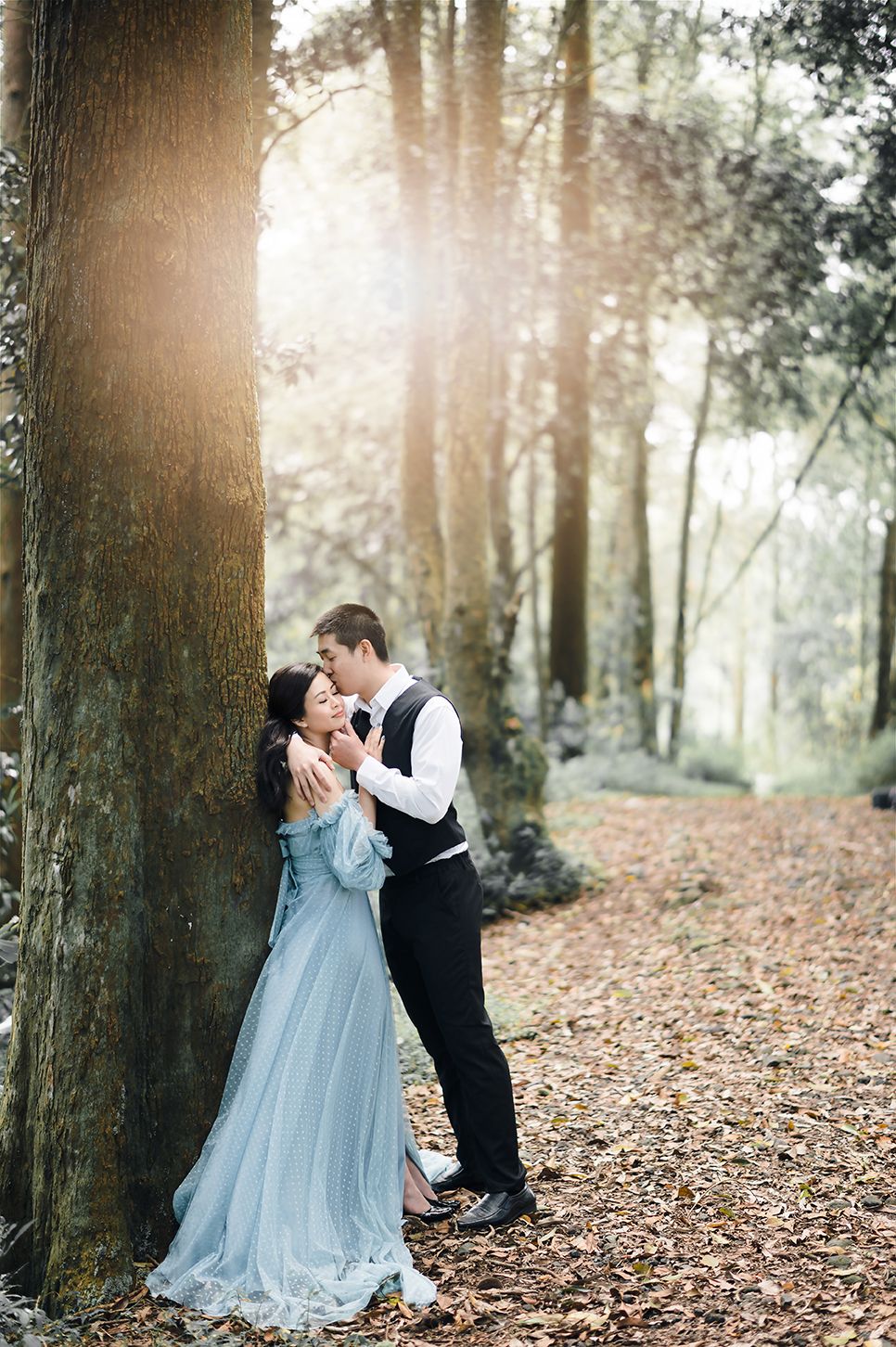 Romantic couple embracing by a large tree in a sunlit forest, woman wearing an elegant blue gown.