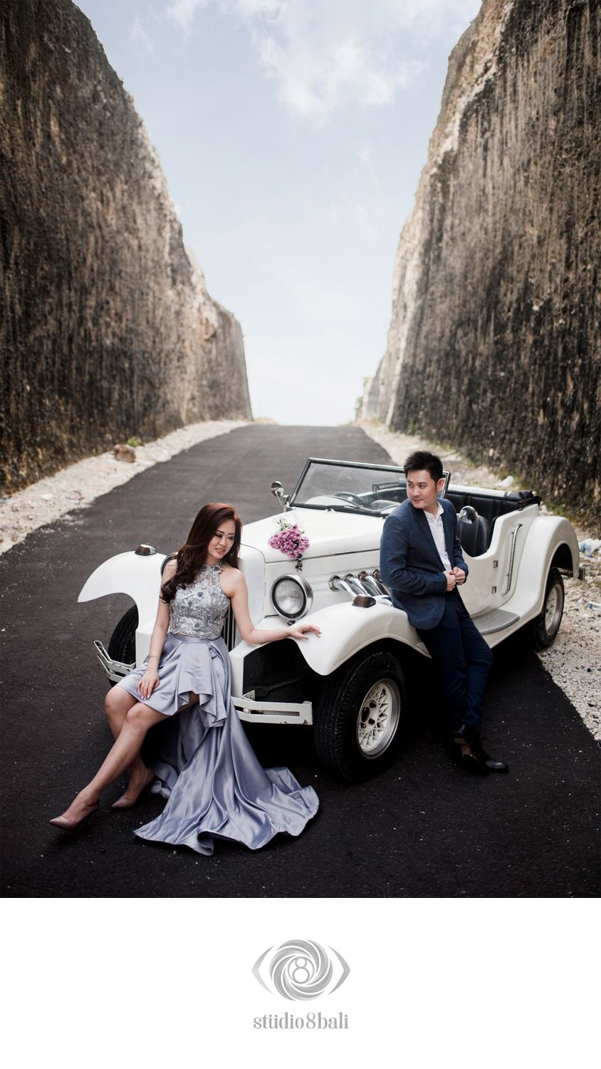 Couple in elegant attire posing with a vintage white convertible car on a scenic road between rocky cliffs in Bali.