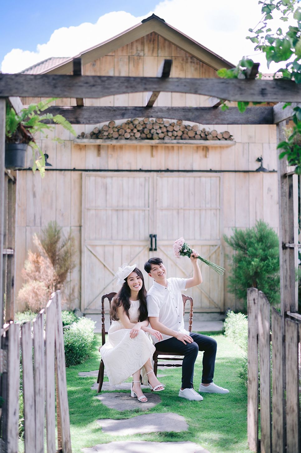 Smiling couple posing on a bench in front of a rustic wooden barn with lush garden surroundings.