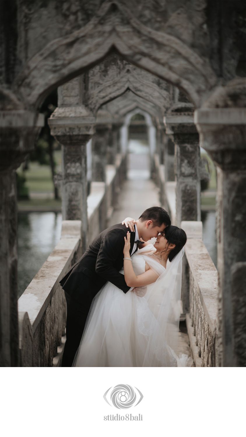 Couple sharing a romantic kiss in wedding attire beneath ornate stone arches at a Bali temple venue.