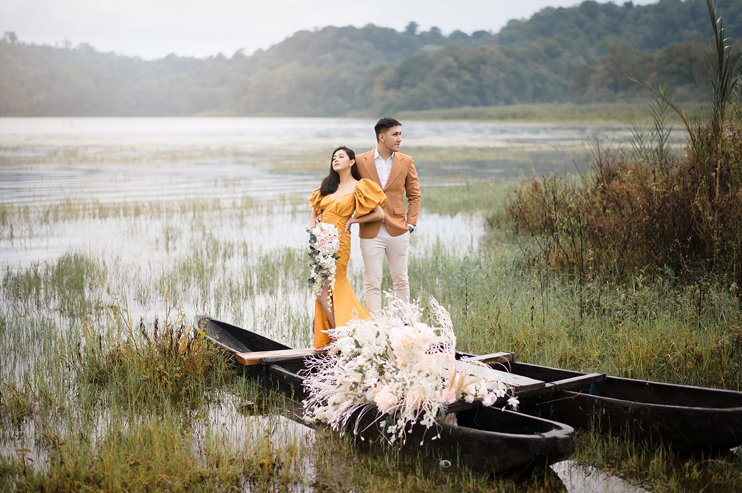 Couple standing in a wooden boat surrounded by flowers on a serene lake during golden hour.