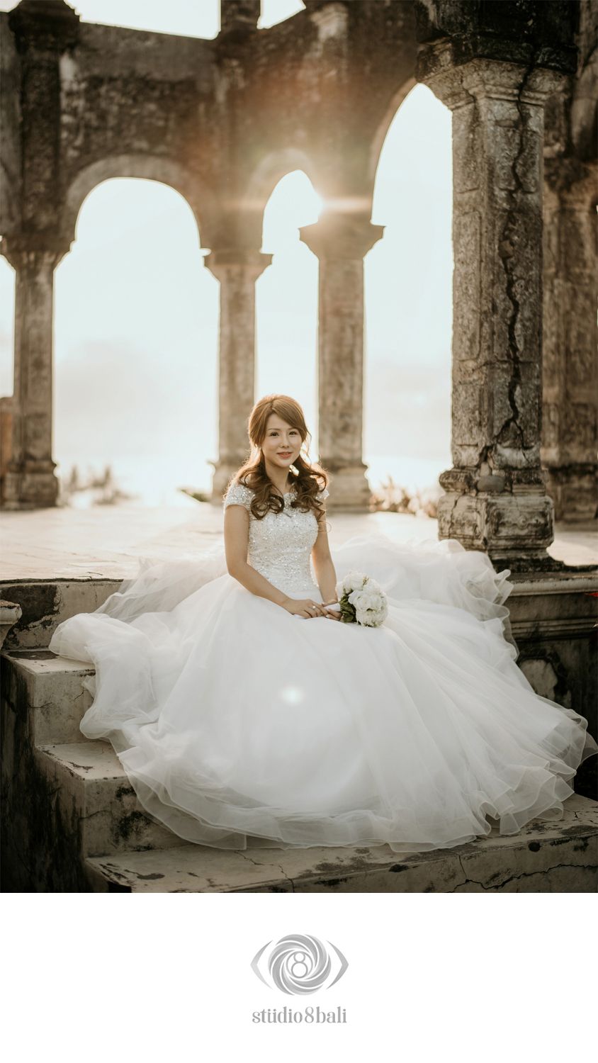 Bride in white ball gown sits on ancient stone steps with sunlit arches and water backdrop, holding white bouquet.