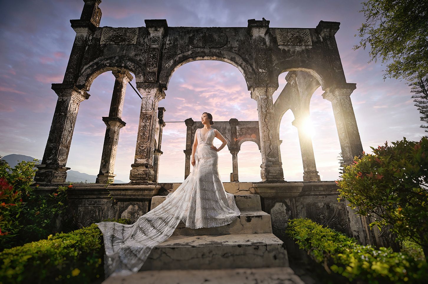 Bride in elegant lace gown poses on ancient stone ruins at sunset with dramatic sky backdrop.