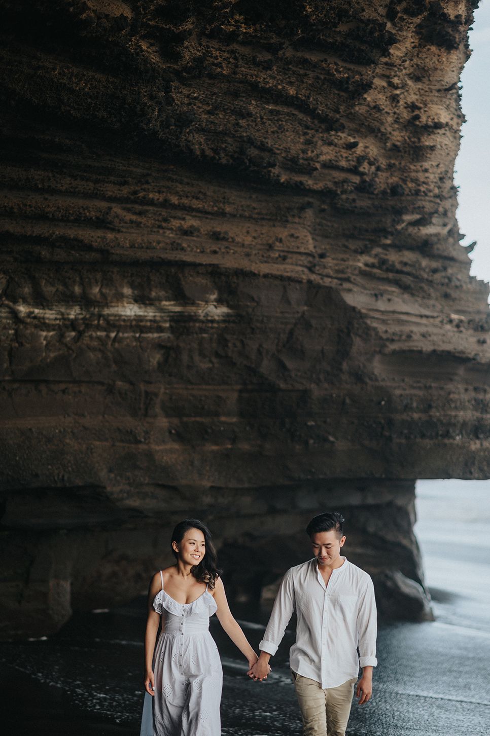 Couple holding hands walking along a dramatic black sand beach beneath towering layered rock cliffs.