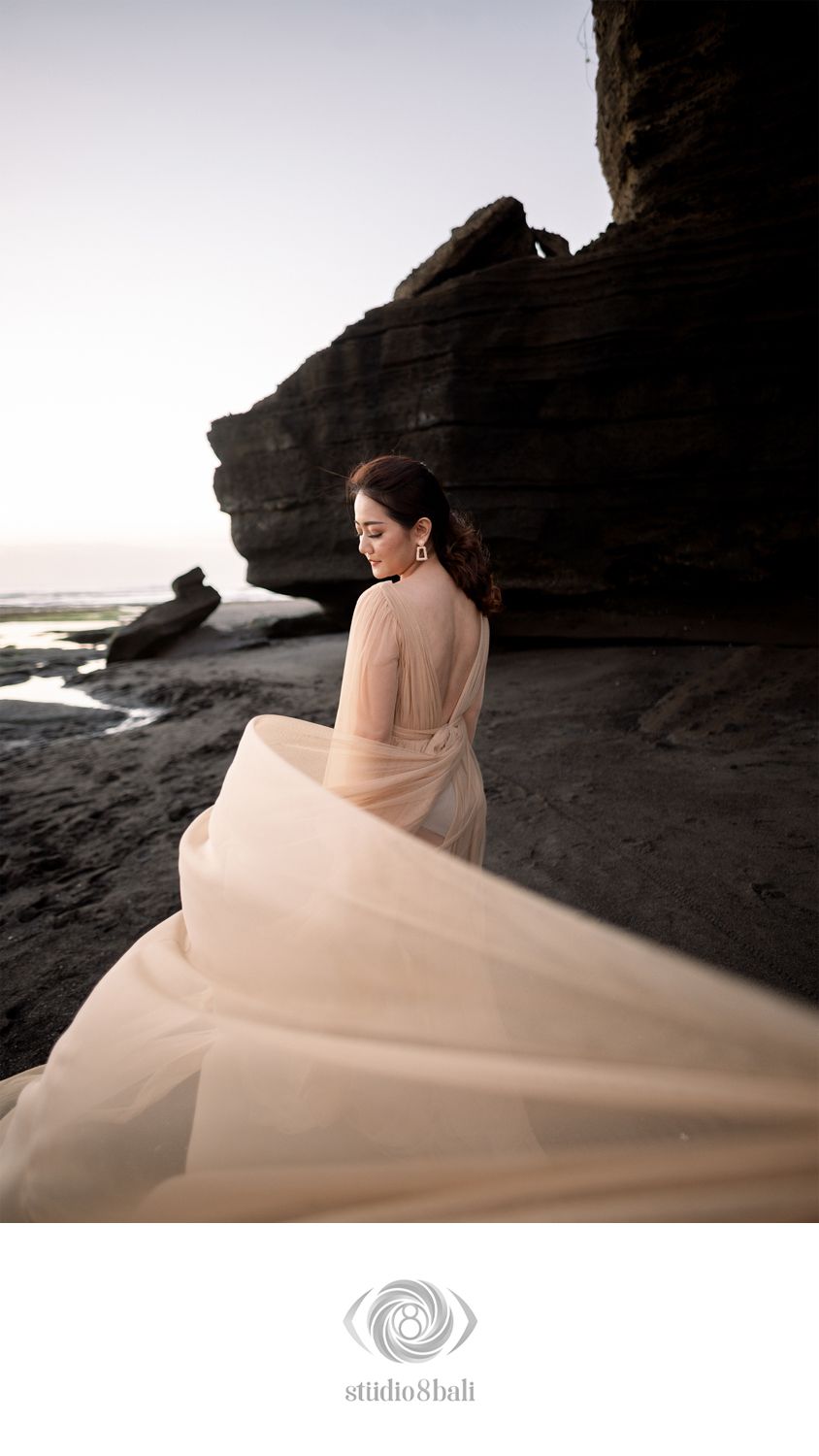 A bride in a flowing sheer gown poses gracefully on a rocky beach at dusk, her dress billowing in the breeze.