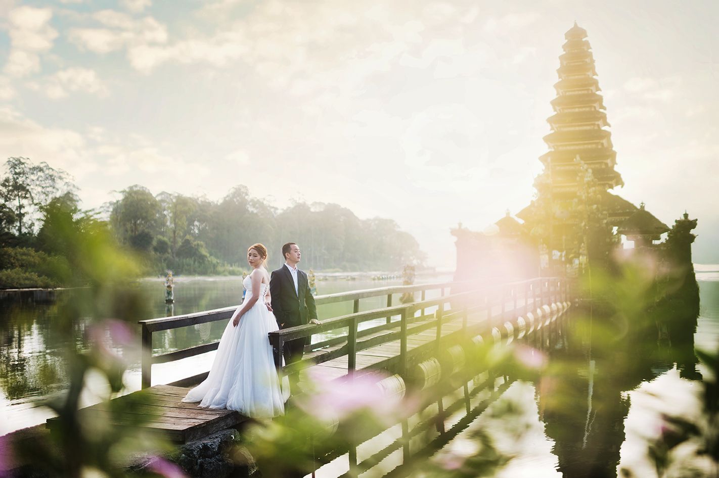 A bride and groom stand on a wooden bridge near a Balinese temple pagoda at sunset, surrounded by lush greenery.