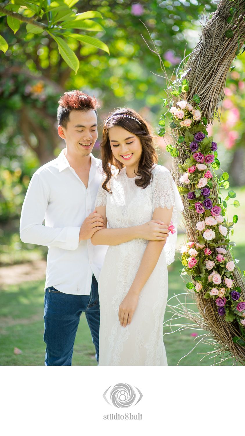 Couple in white attire poses by a floral-adorned tree arch in a lush garden for a romantic pre-wedding shoot.