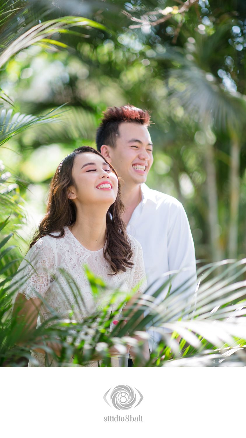 A smiling couple poses among lush tropical palm leaves in a bright outdoor engagement photo shoot.