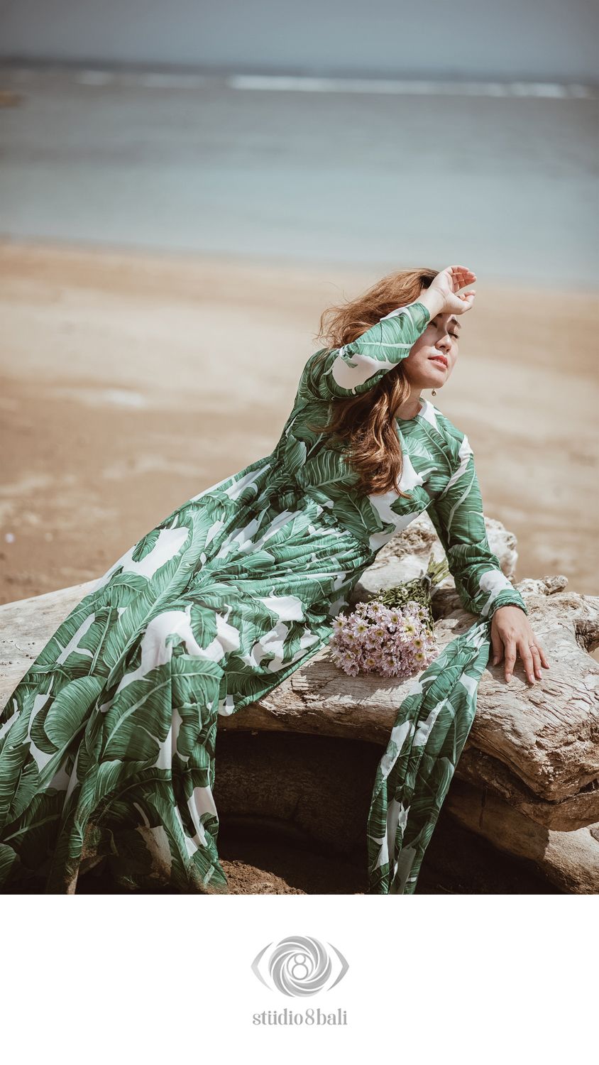 Woman in green tropical print maxi dress poses on driftwood at beach holding wildflowers.