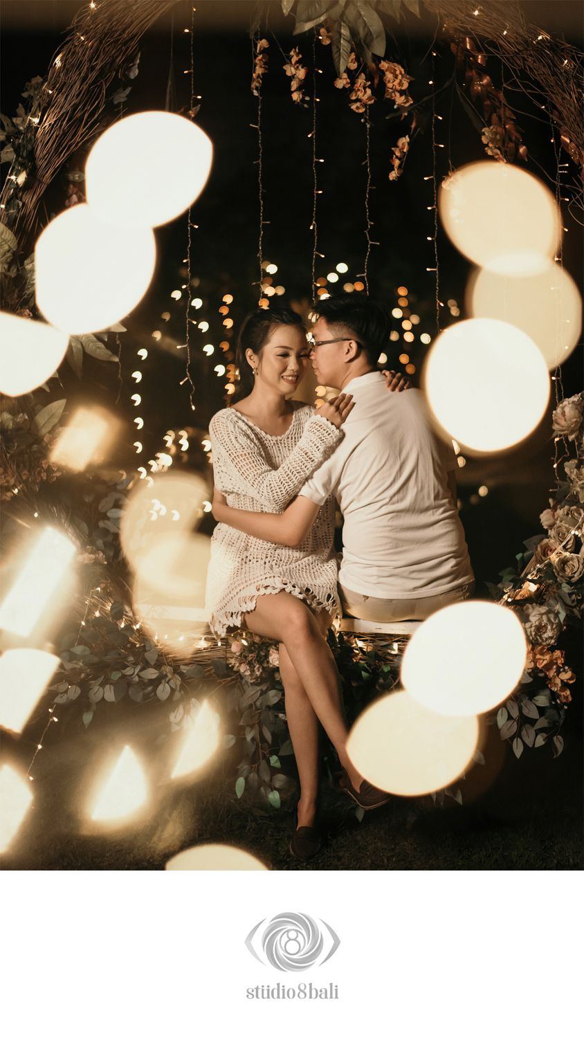 Romantic couple sitting on a swing surrounded by bokeh lights and fairy string lights in a dreamy outdoor setting.
