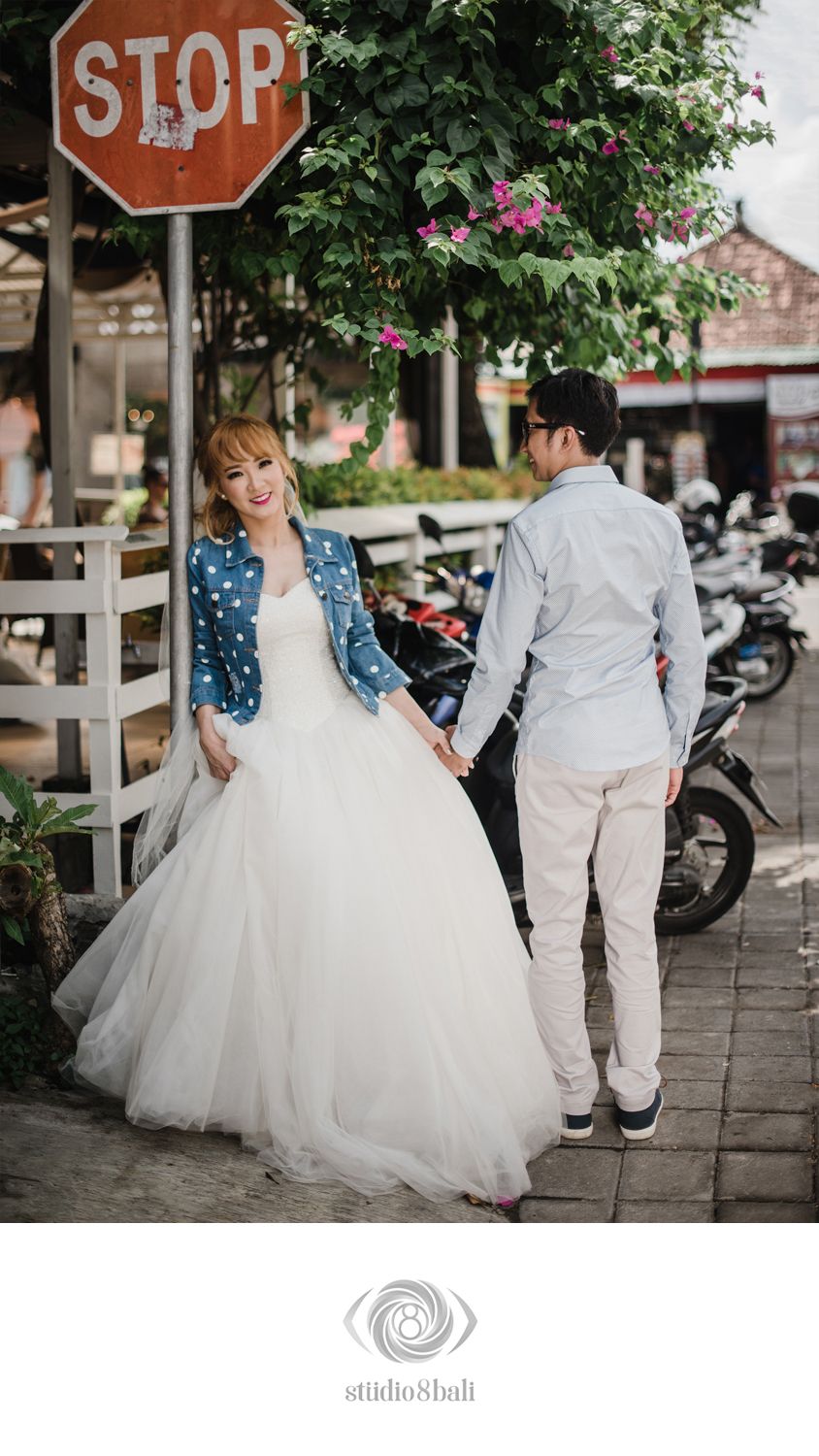 Couple posing near a stop sign, bride in white gown and denim jacket holding hands with groom in casual attire outdoors.