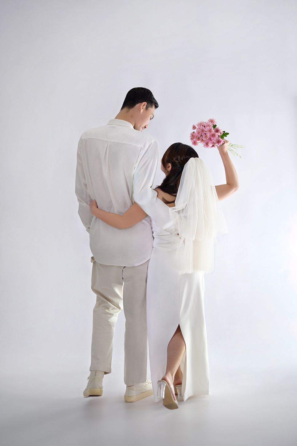 Bride in white dress holding pink flowers stands with groom in white outfit, viewed from behind on white background.
