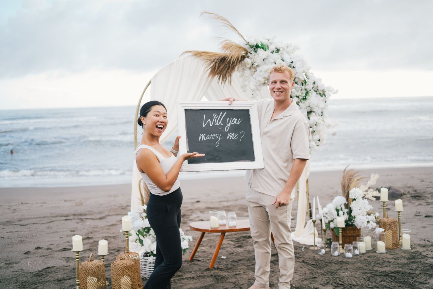 Couple on beach with will you marry me chalkboard sign, floral arch, candles, and pampas grass decor at sunset.