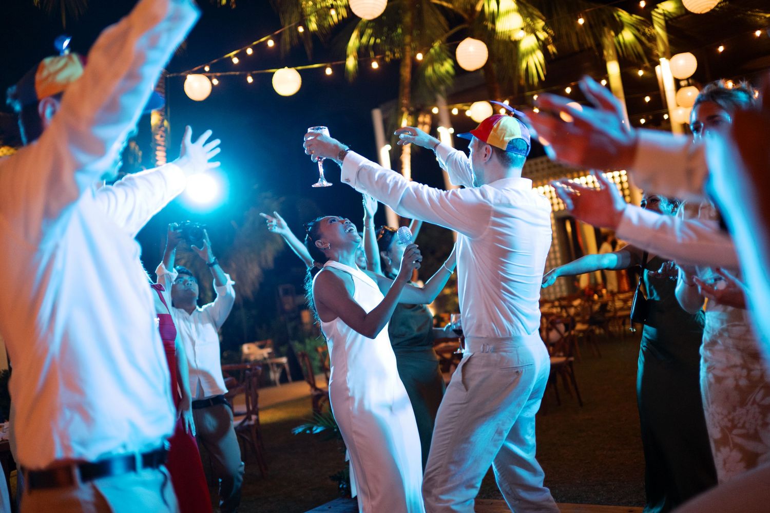 Guests dancing joyfully at an outdoor wedding reception at night with string lights and lanterns.