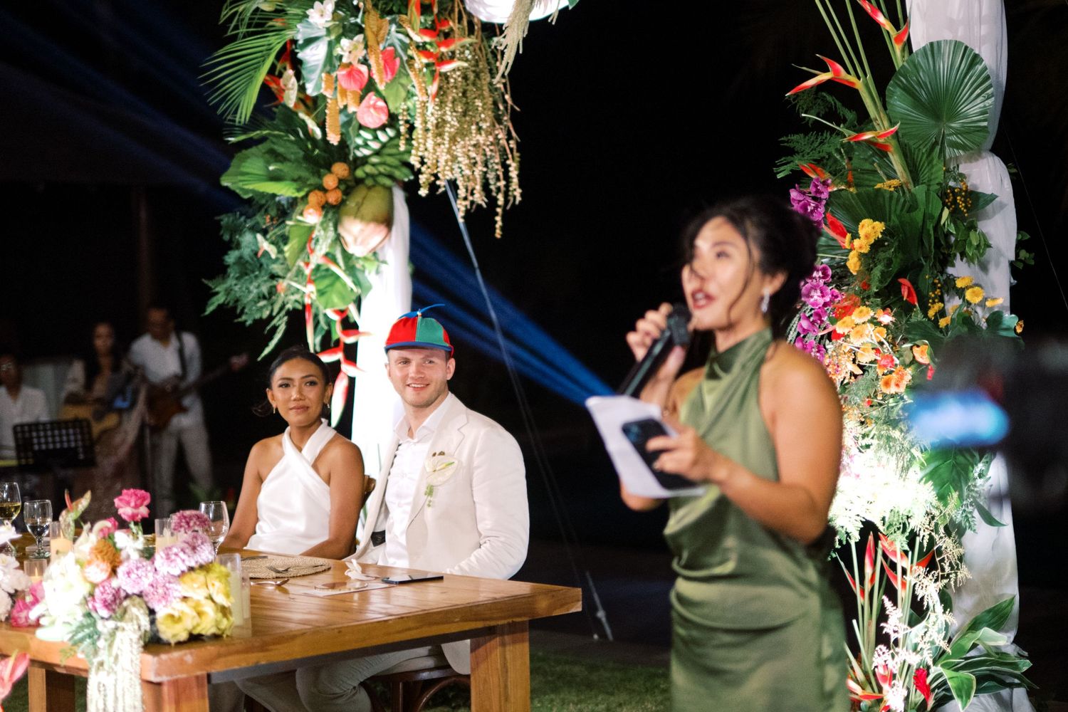 A woman in a green dress speaks into a microphone at an outdoor tropical wedding reception while the couple sits at a decorated table.