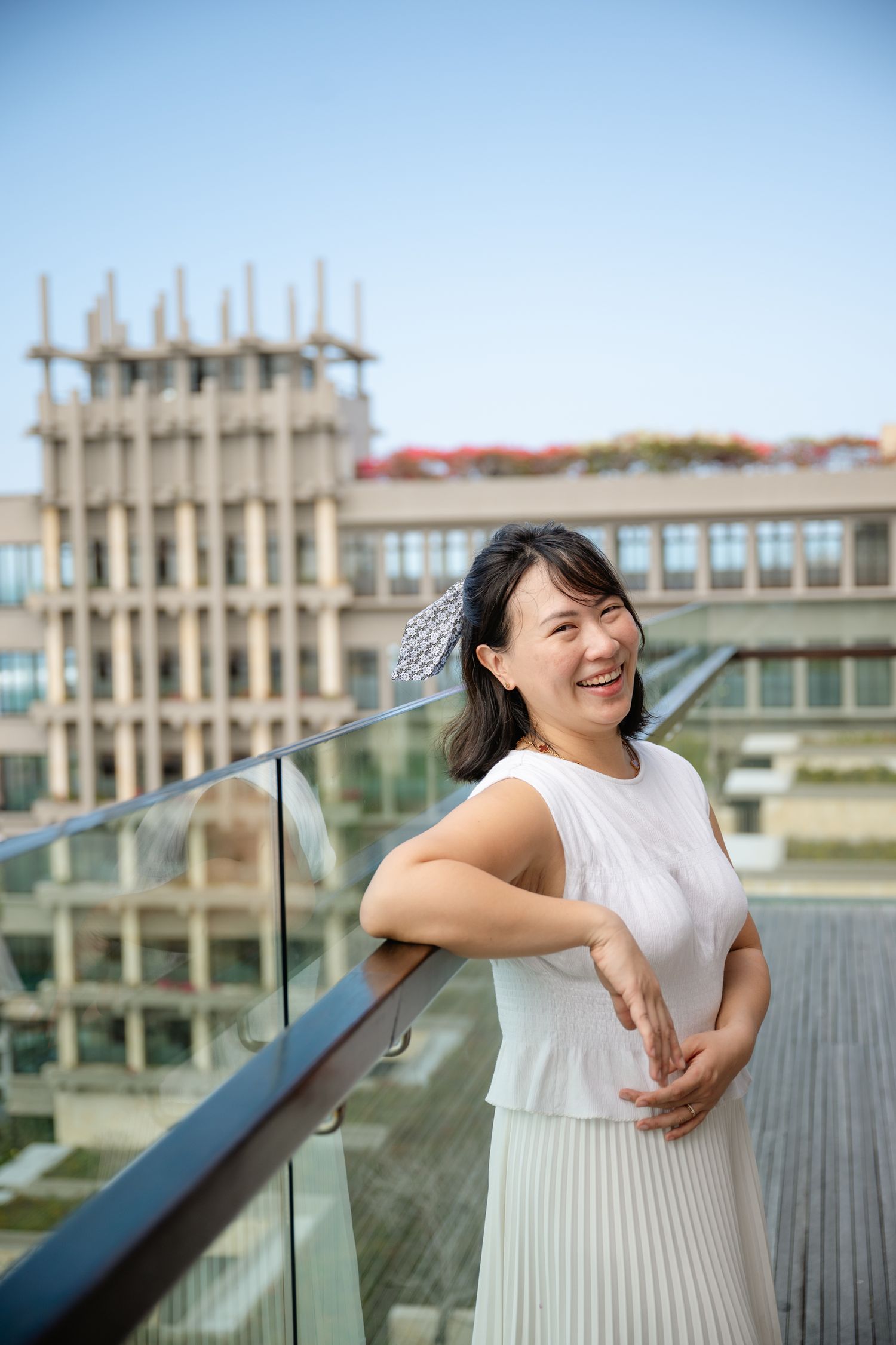 Smiling Asian woman in white outfit leaning on glass balcony railing with urban buildings in background.
