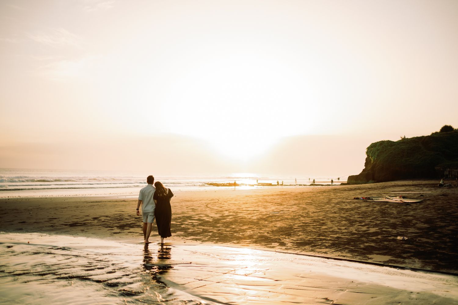 Couple standing together on a sandy beach at golden sunset with ocean waves and rocky cliffs in the background.
