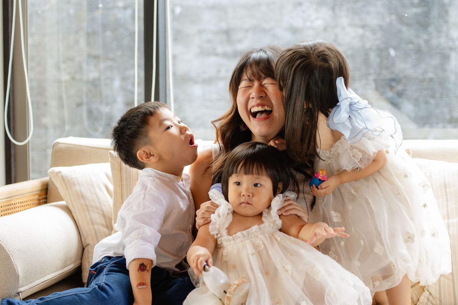 A joyful Asian mother laughs with her three young children on a couch near a bright window.