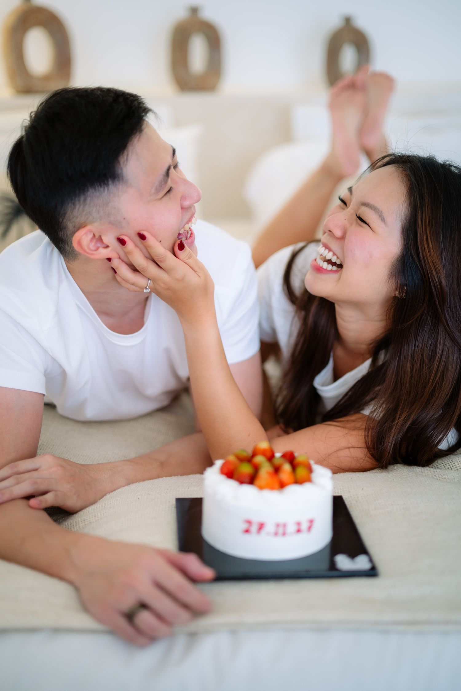 Happy Asian couple lying on bed, laughing and sharing a strawberry cake with a special date, celebrating anniversary.