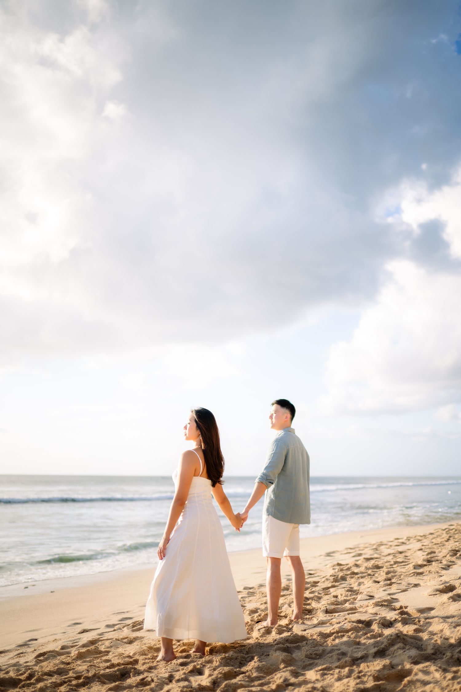 A couple holds hands on a sandy beach, woman in white dress, man in casual attire, with ocean waves and cloudy sky behind.
