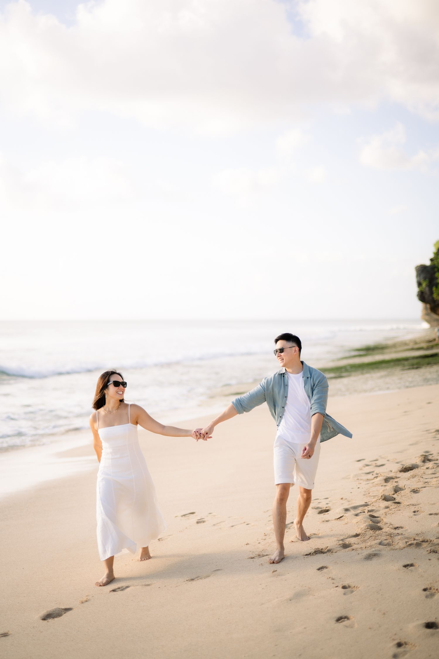 A couple walks hand-in-hand on a sandy beach, wearing white outfits and sunglasses, with ocean waves in the background.
