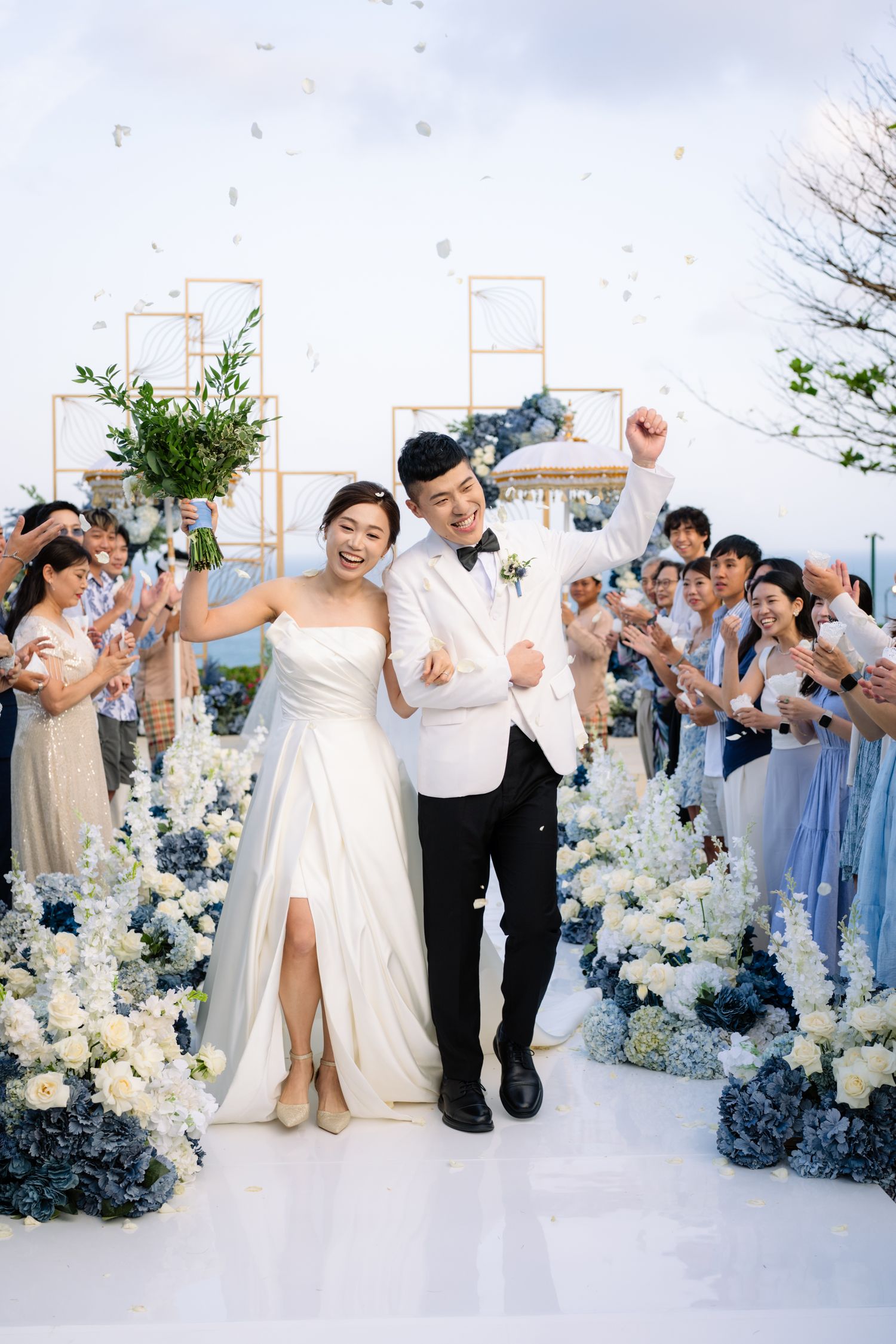 Joyful bride in a strapless white gown and groom in a white tuxedo walk the aisle as guests cheer at an oceanside wedding ceremony.