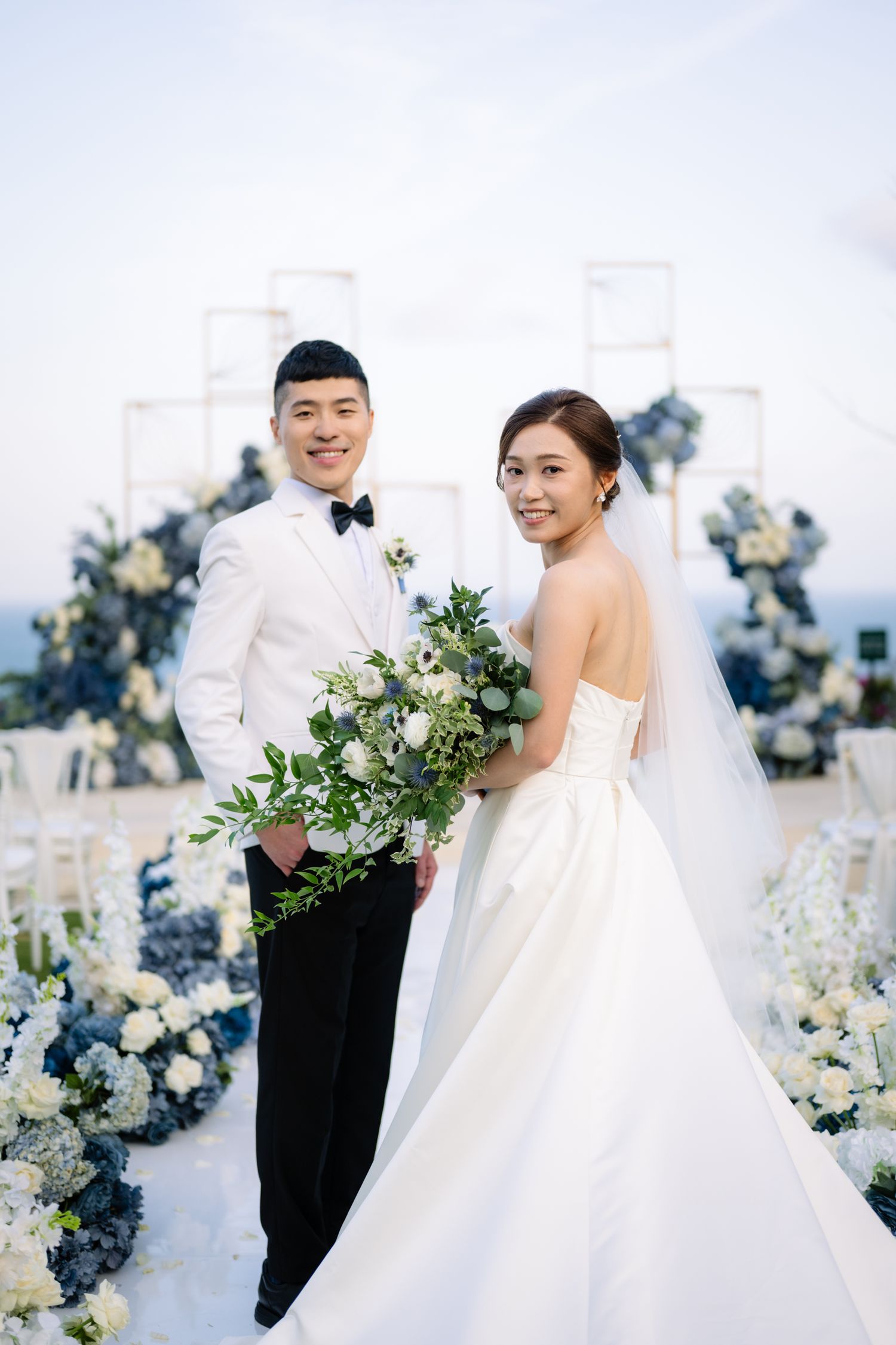 Bride in white gown and veil holding a green bouquet beside a smiling groom in white jacket at an oceanside wedding with blue floral decor.