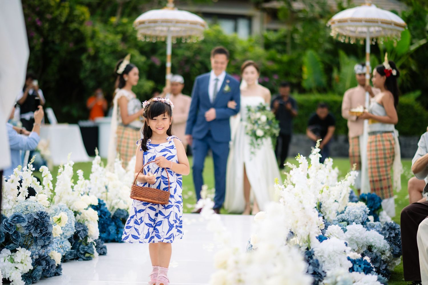 Flower girl in a blue floral dress walks down an outdoor wedding aisle lined with white and blue flowers as the bride and groom follow.