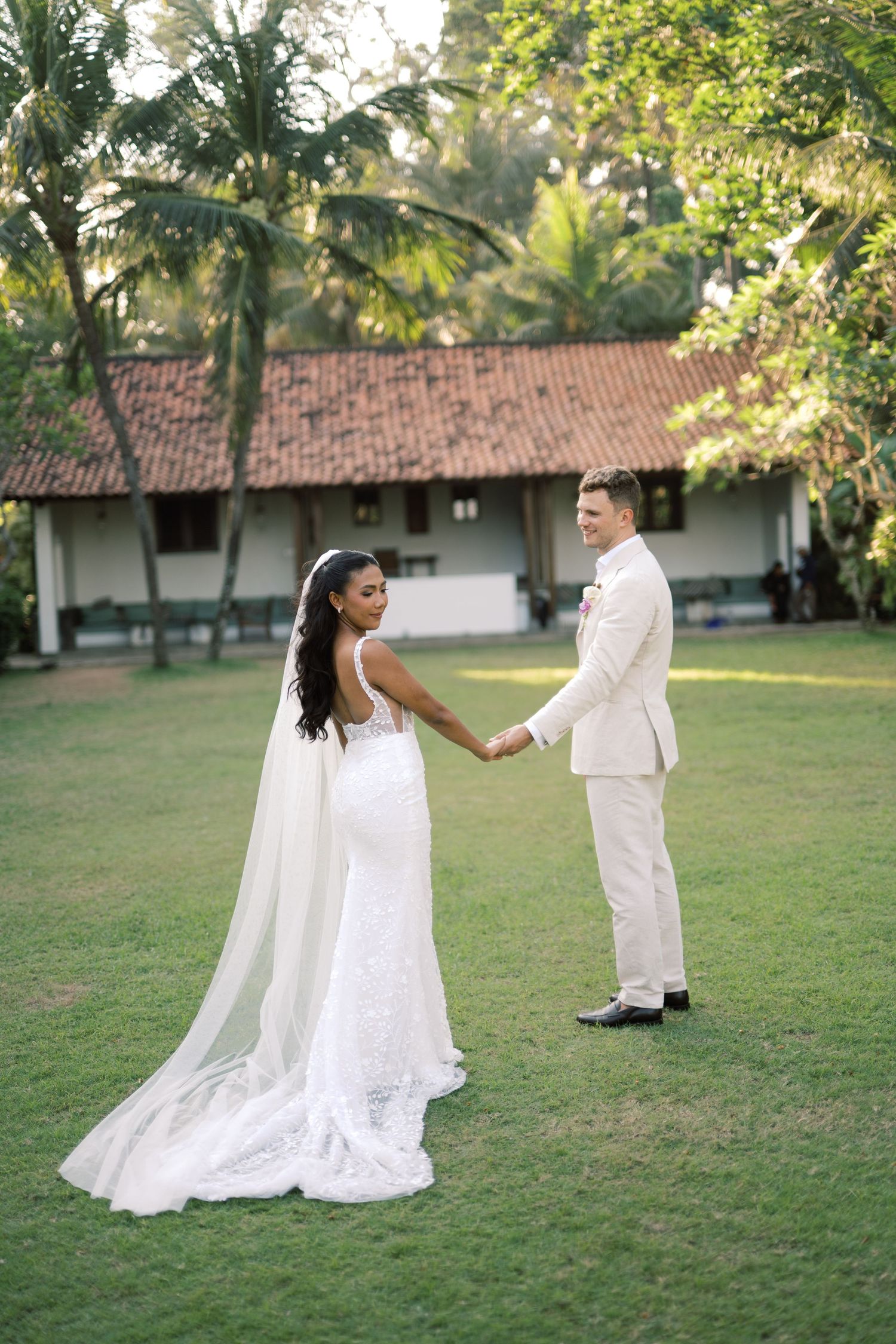 Bride in white lace gown with cape veil holds hands with groom in cream suit on lush green lawn.