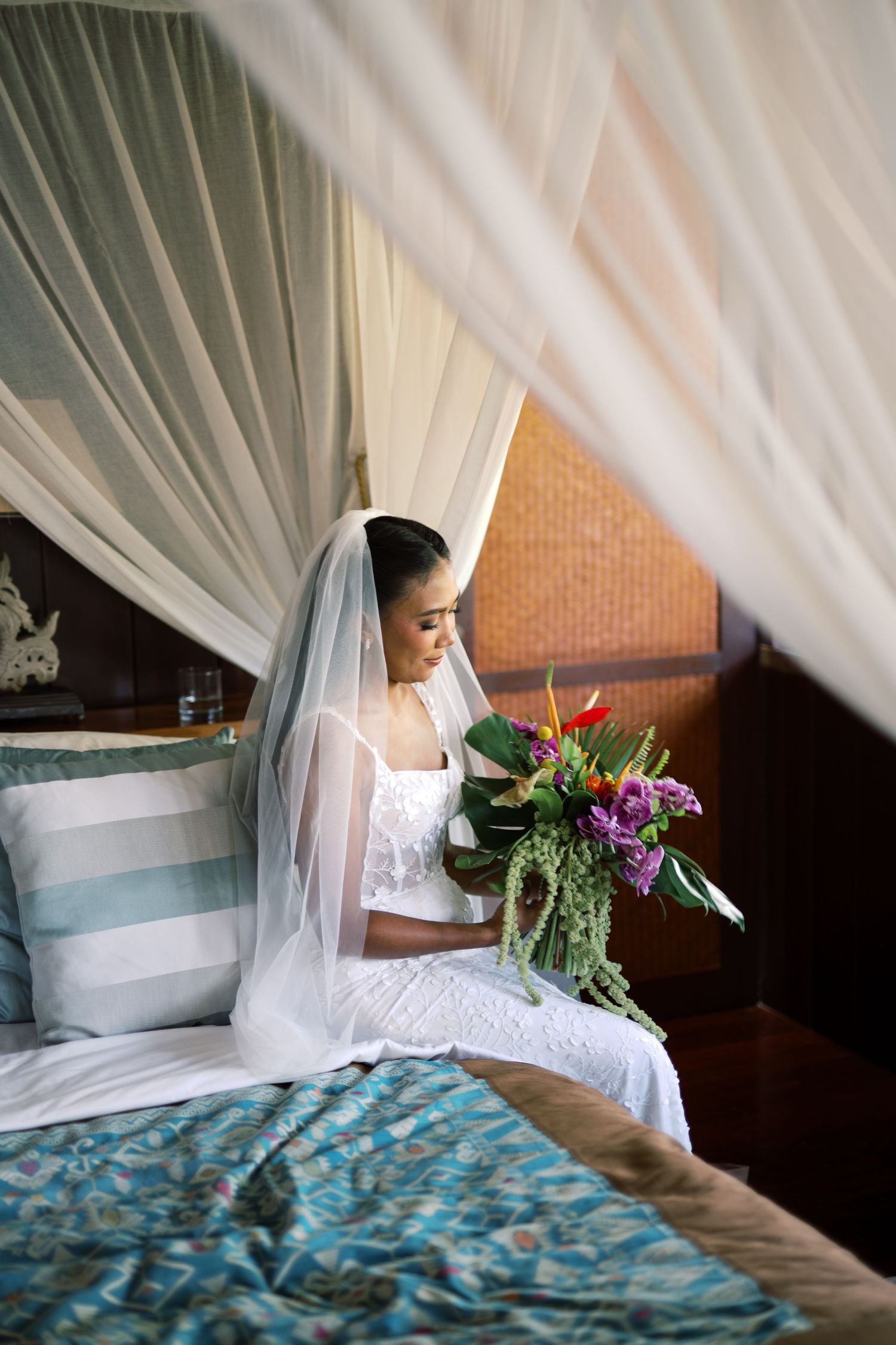 Bride in white lace gown and veil sits on canopy bed holding tropical bouquet.