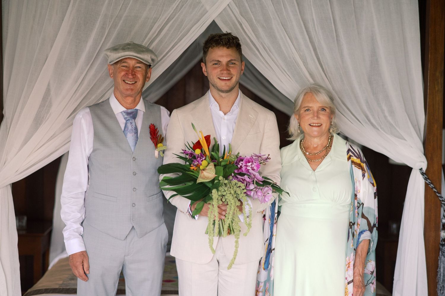 Three people posing together at an outdoor wedding ceremony under white drapes, man in center holding tropical bouquet.