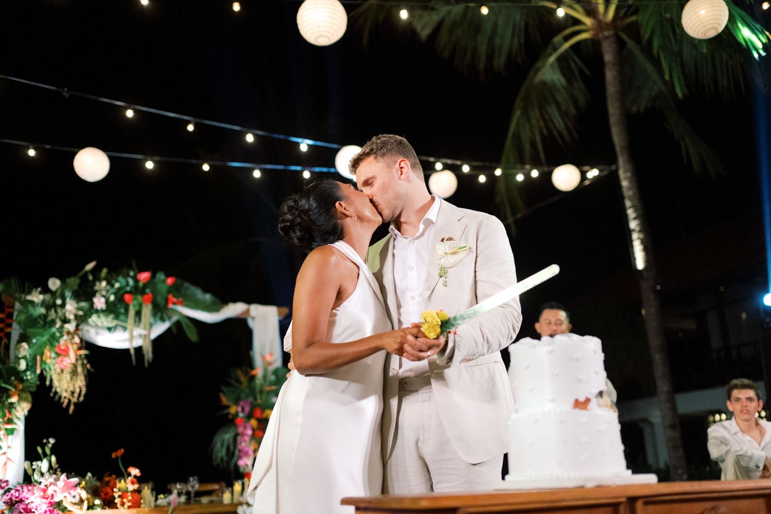 Bride and groom share a romantic kiss while cutting their wedding cake at a tropical outdoor reception.