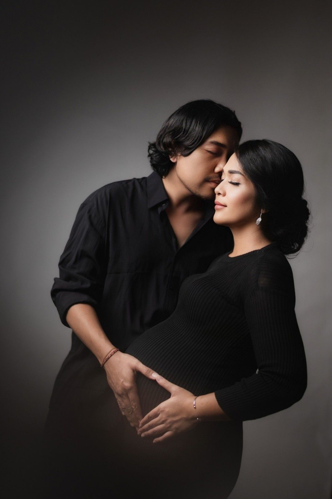 A couple in black outfits poses tenderly, man kissing womans temple while cradling her pregnant belly in a studio.