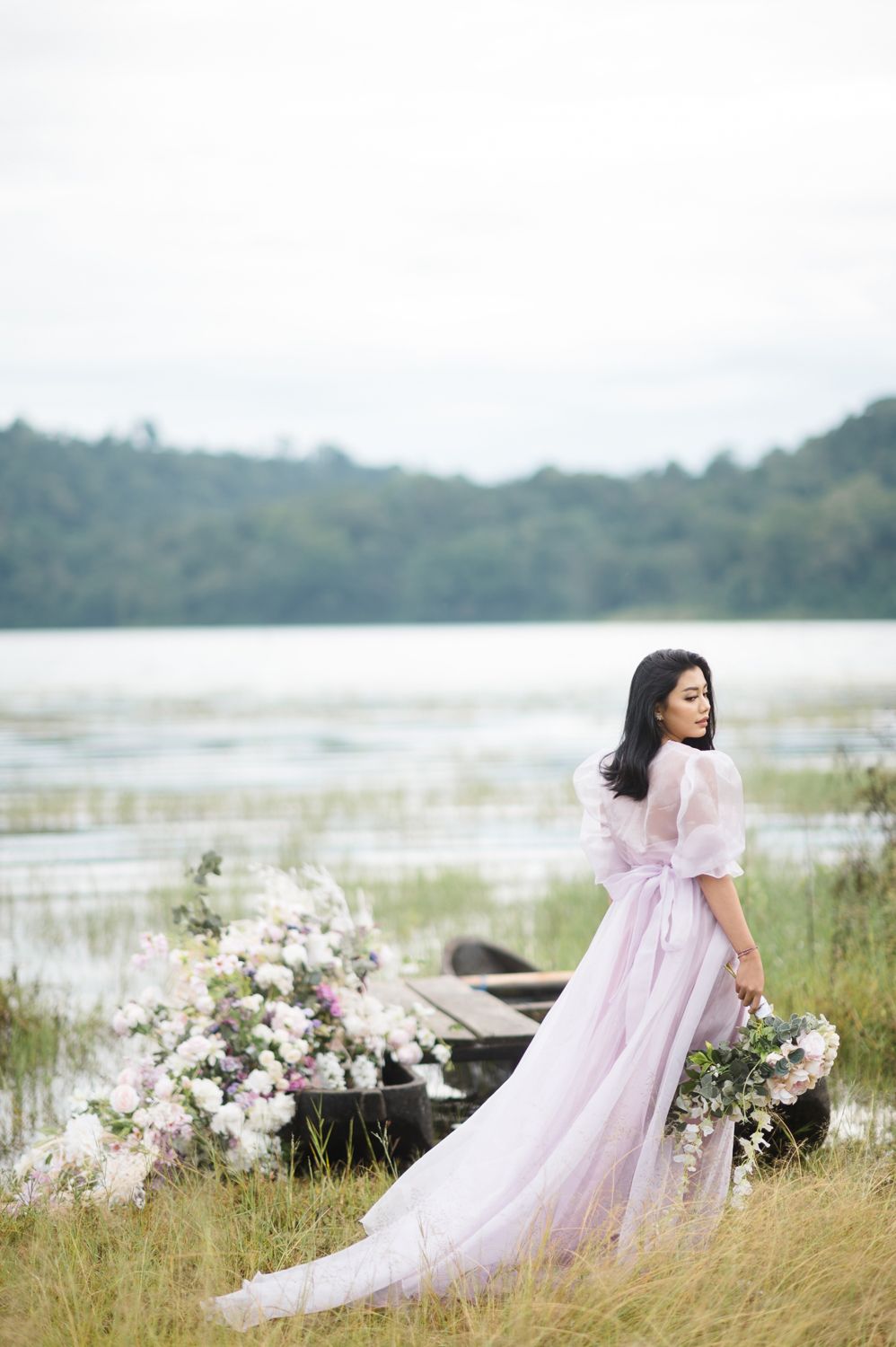 Woman in lavender gown holds flowers by a lakeside dock with lush greenery and floral arrangements in soft light.
