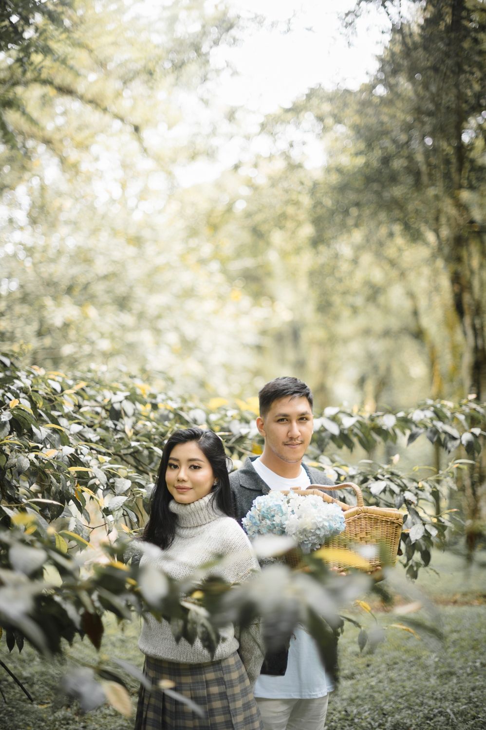 Couple posing in a lush green forest, man holding a wicker basket with blue flowers, woman in cozy turtleneck sweater.