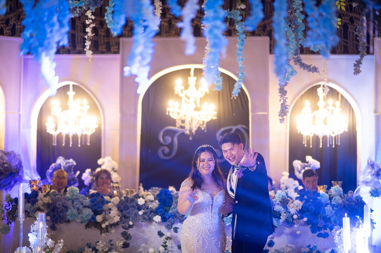 Bride and groom smiling at elegant wedding reception with blue floral decor, chandeliers, and romantic lighting.