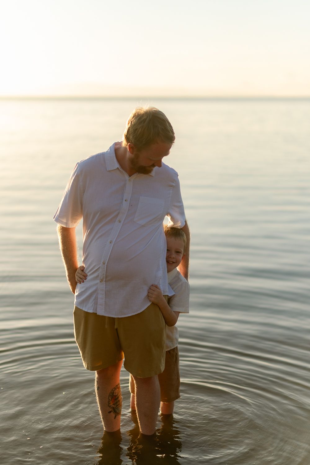 A father and young son playing together in shallow ocean water during a warm golden sunset.