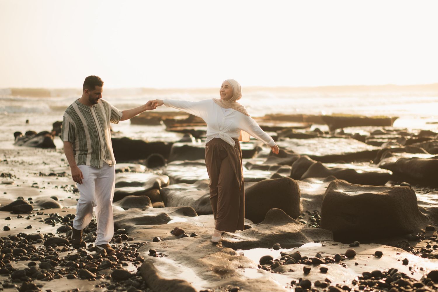 Couple walking on rocky beach at sunset, woman balancing with arms outstretched, golden light reflecting on wet stones.