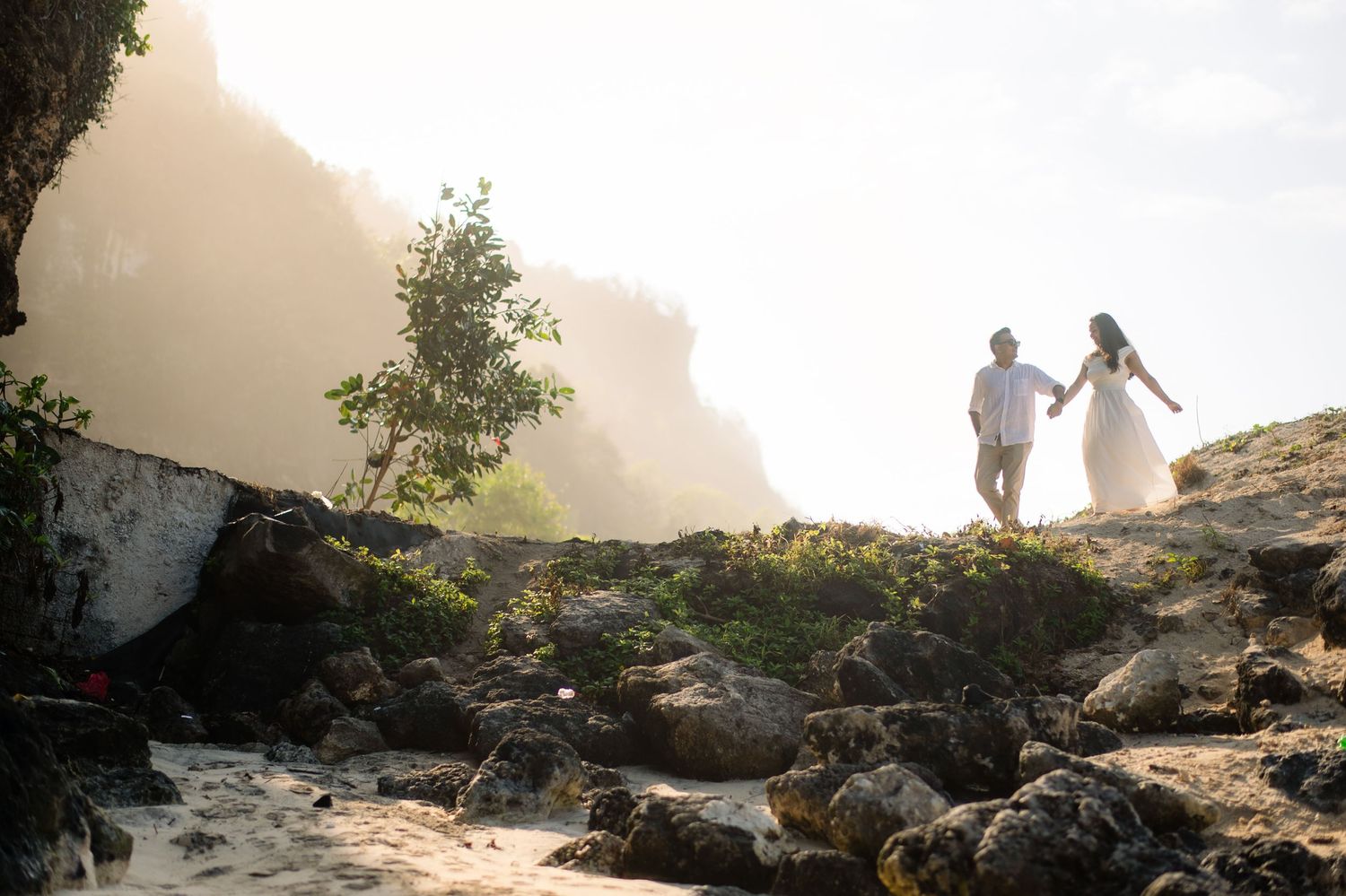 Couple holding hands walking on rocky beach with misty cliffs and lush greenery in background during golden hour.