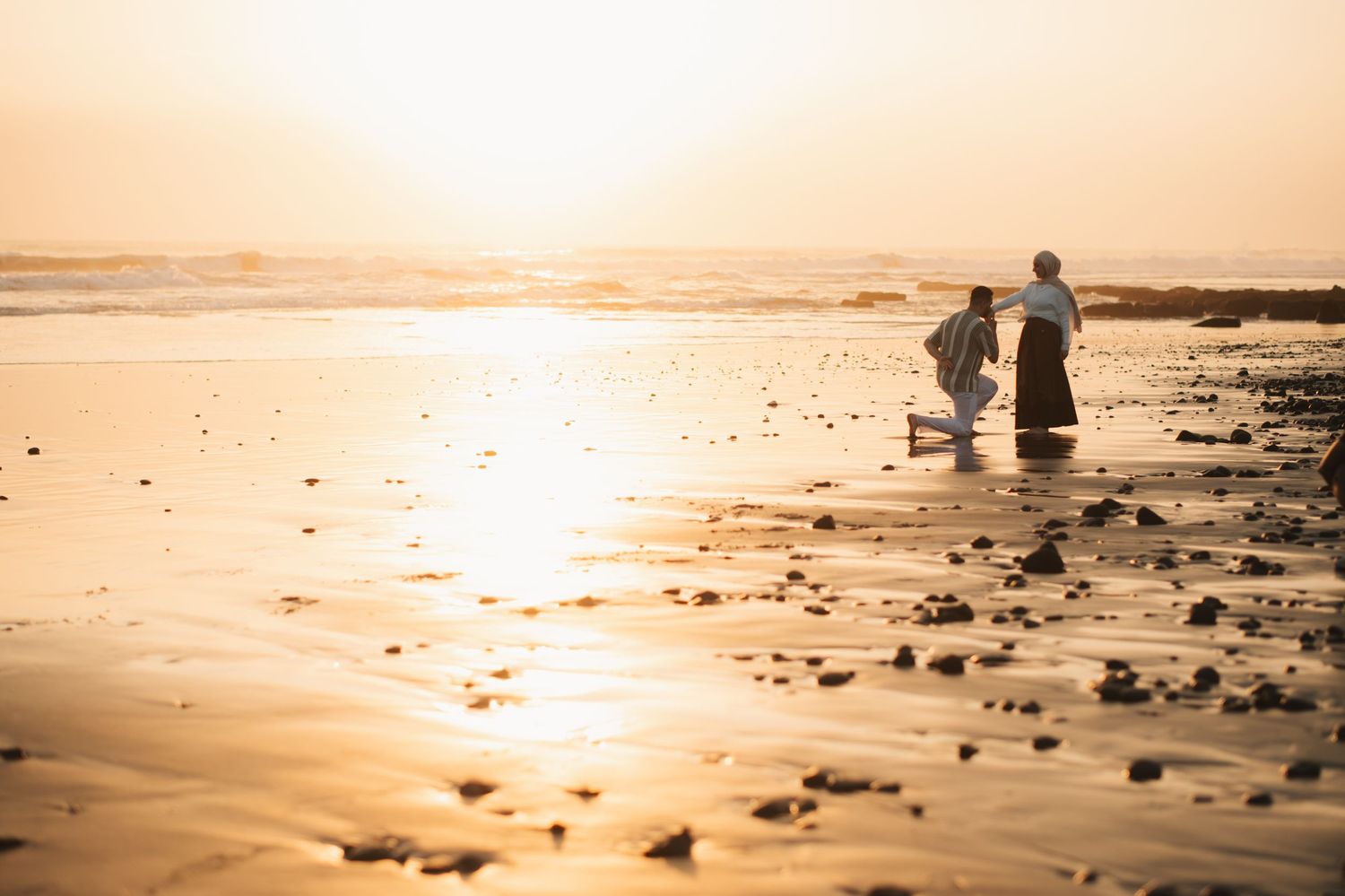 Two people walk along a golden sandy beach at sunset, with waves reflecting warm sunlight in a serene coastal scene.