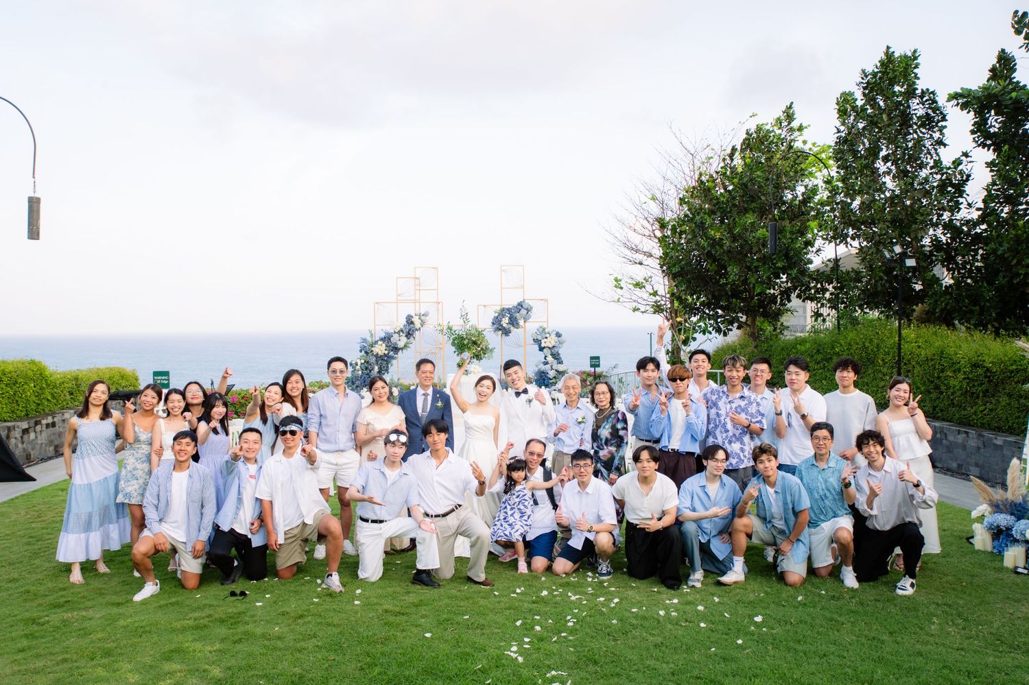 Large wedding party in blue and white attire posing together on a grassy lawn with a floral arch and ocean view in the background.