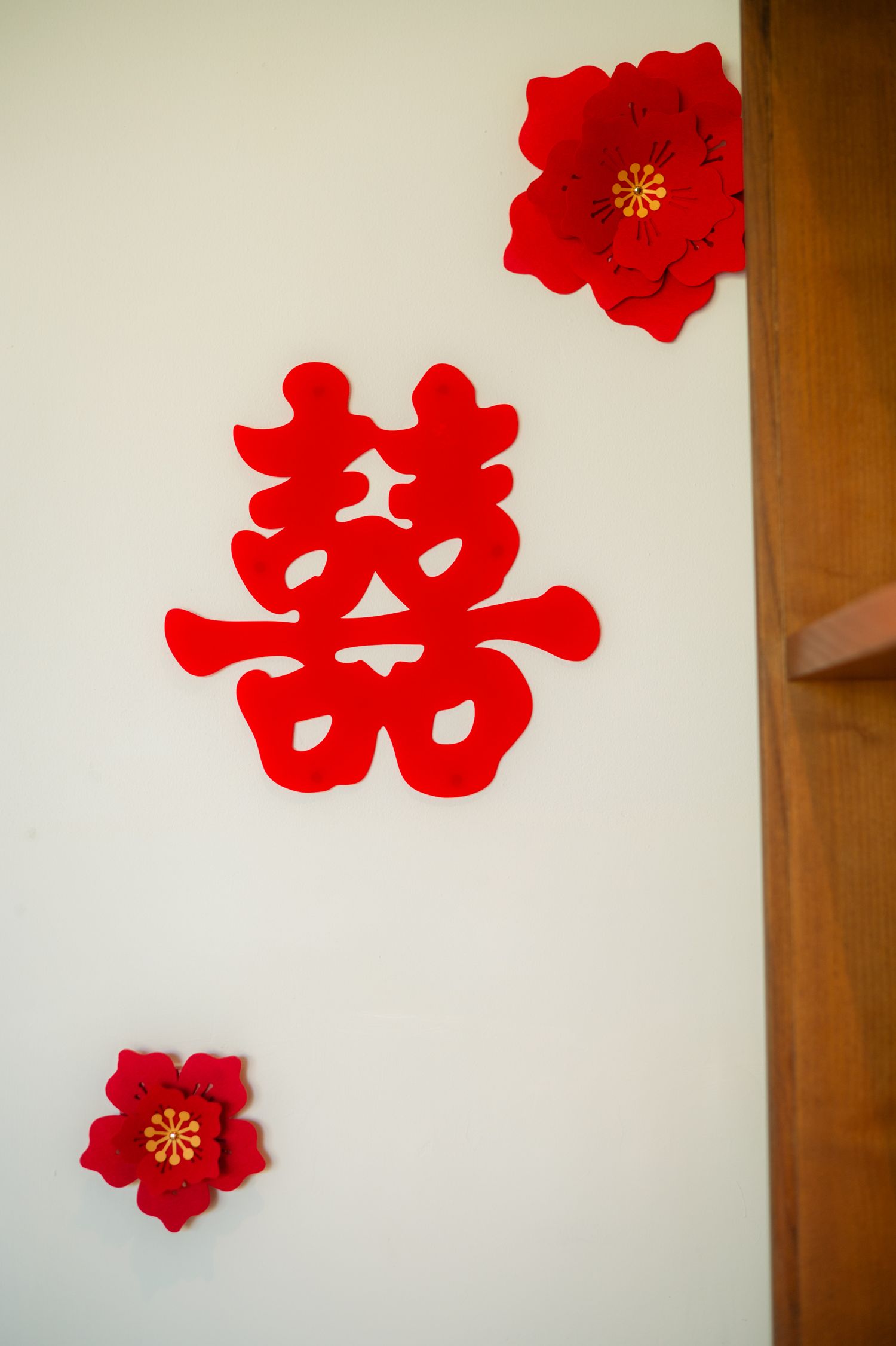Red paper double happiness xi symbol with two red paper flowers decorating a white wall beside a wooden door frame for a Chinese wedding.