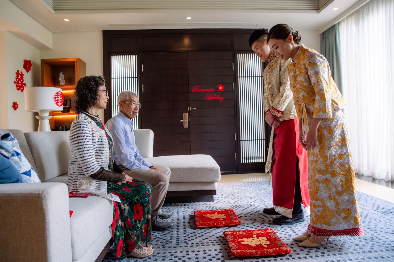 Young couple in traditional Chinese wedding attire kneel at red cushions to serve tea to seated elderly parents in a bright living room.