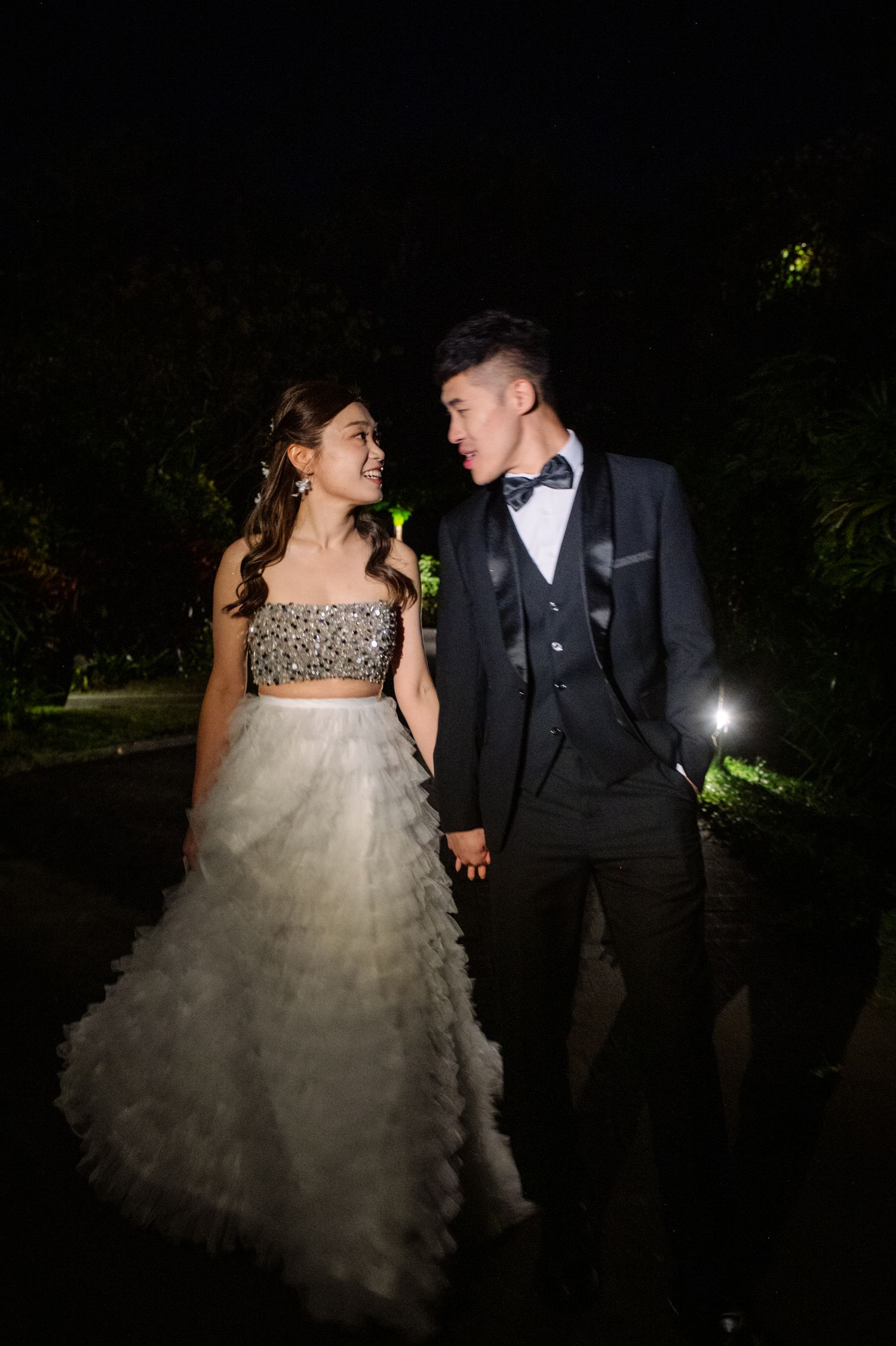 A smiling couple in formal evening attire holds hands and gazes at each other while walking outdoors at night.